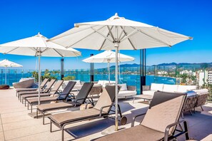 Eine Dachterrasse mit Liegestühlen und Sonnenschirmen, mit Blick auf eine Stadtlandschaft und ein Gewässer.