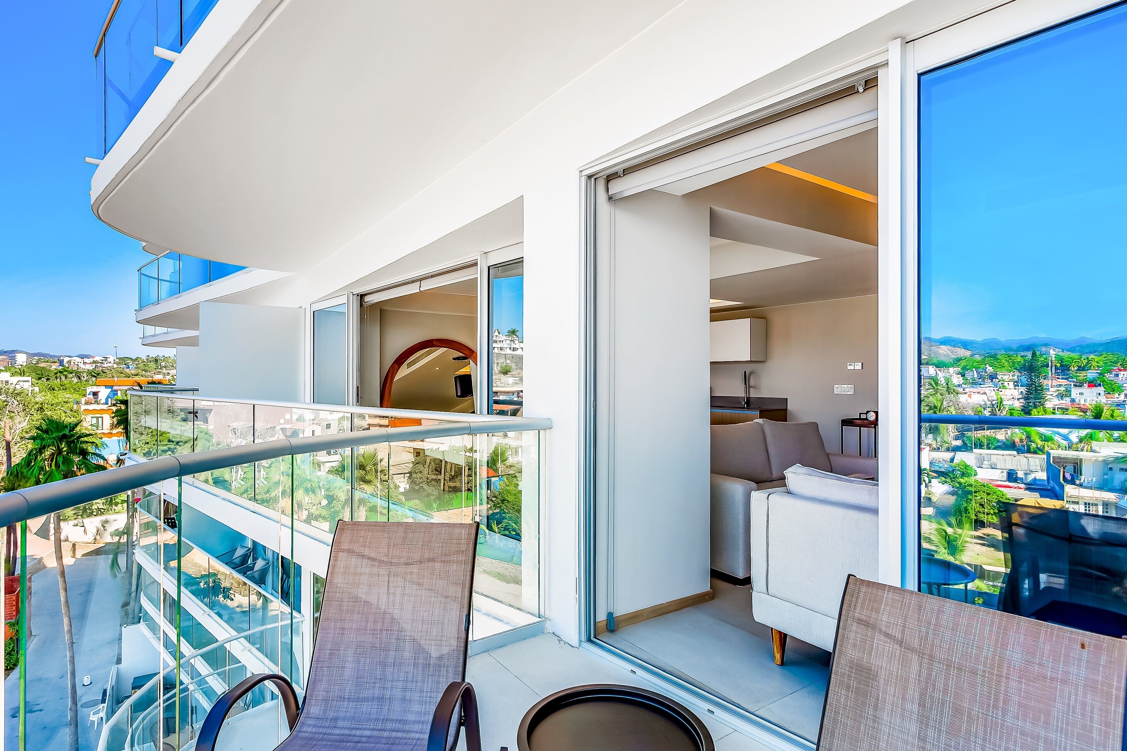 En moderne balkon med glasgelændere, en sofa og et spiseområde med udsigt over byen.