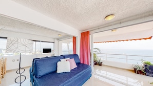 A modern living room with a blue sofa, a white fluffy pillow, and a pink pillow. There is a large window with a view of the sea. - Central Puerto Vallarta Condo With Pool, Ocean Views & Fast Wifi - Walk to Beach (Puerto Vallarta)