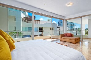 Ein modernes Hotelzimmer mit einem großen Bett, einer Couch und einem Balkon mit Blick auf andere Gebäude und Palmen.