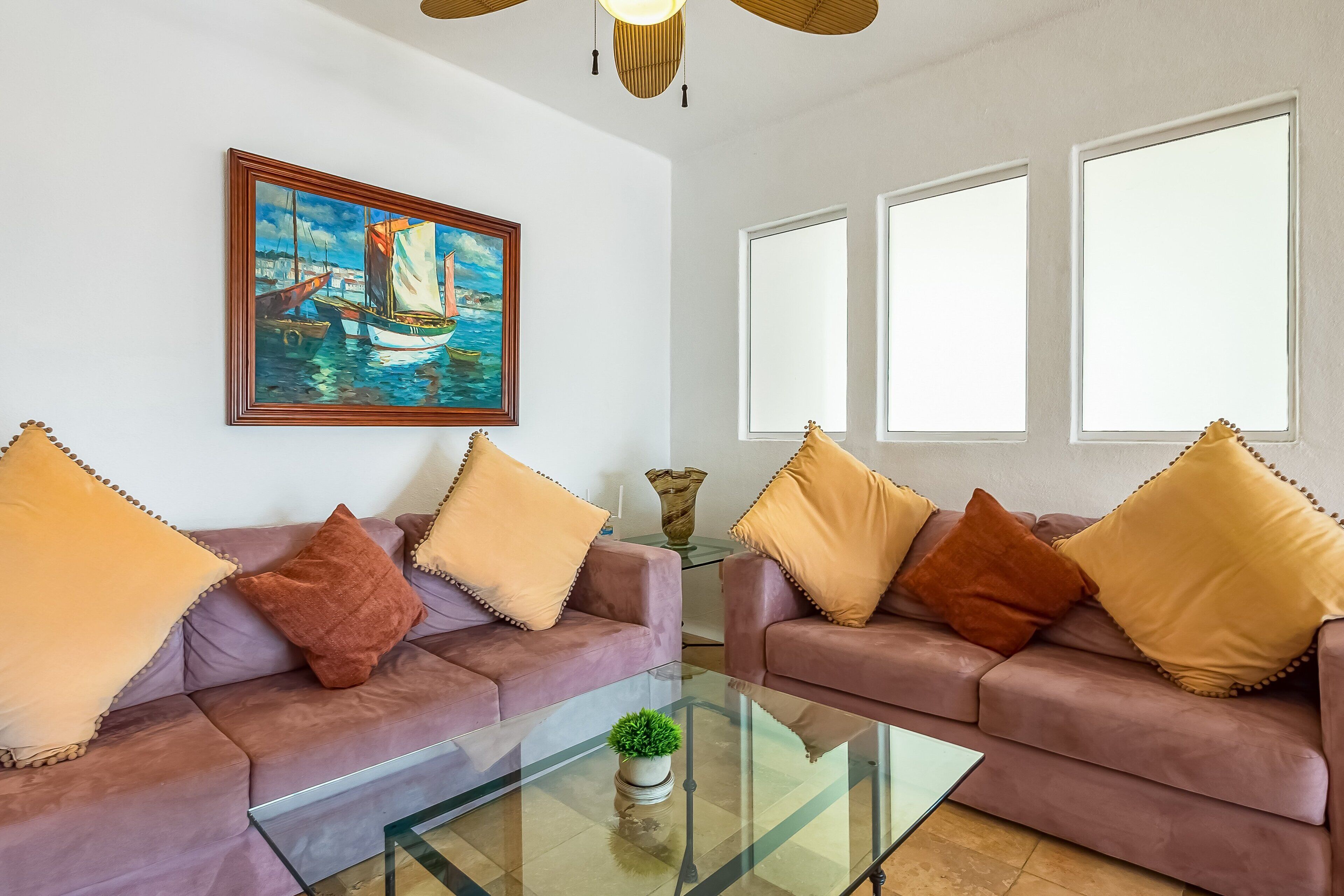 Een woonkamer met twee banken, een glazen salontafel, een schilderij van een zeilboot, en drie ramen.