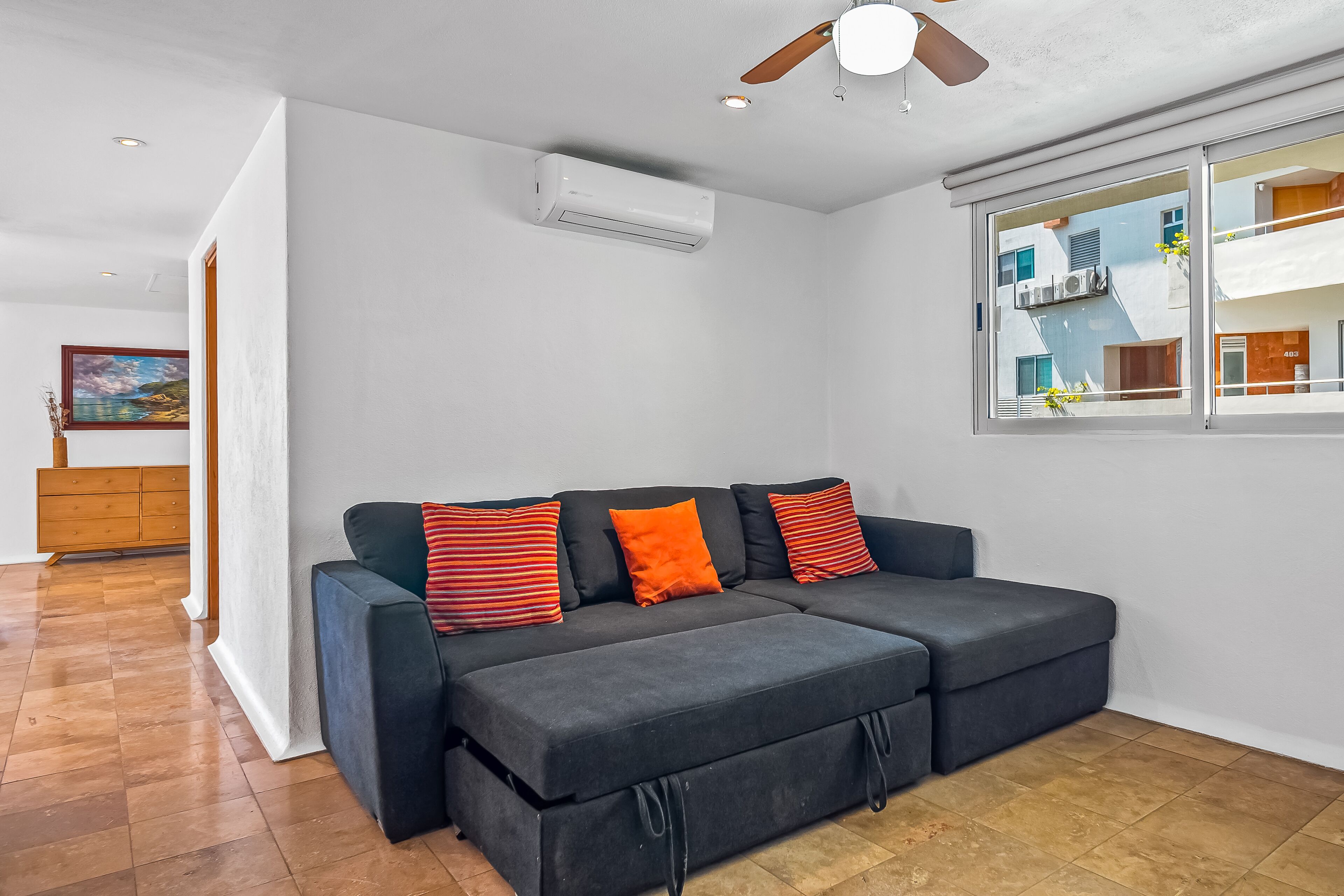 Un salon moderne avec un canapé d’angle gris, un ventilateur de plafond et une grande fenêtre donnant sur des immeubles.