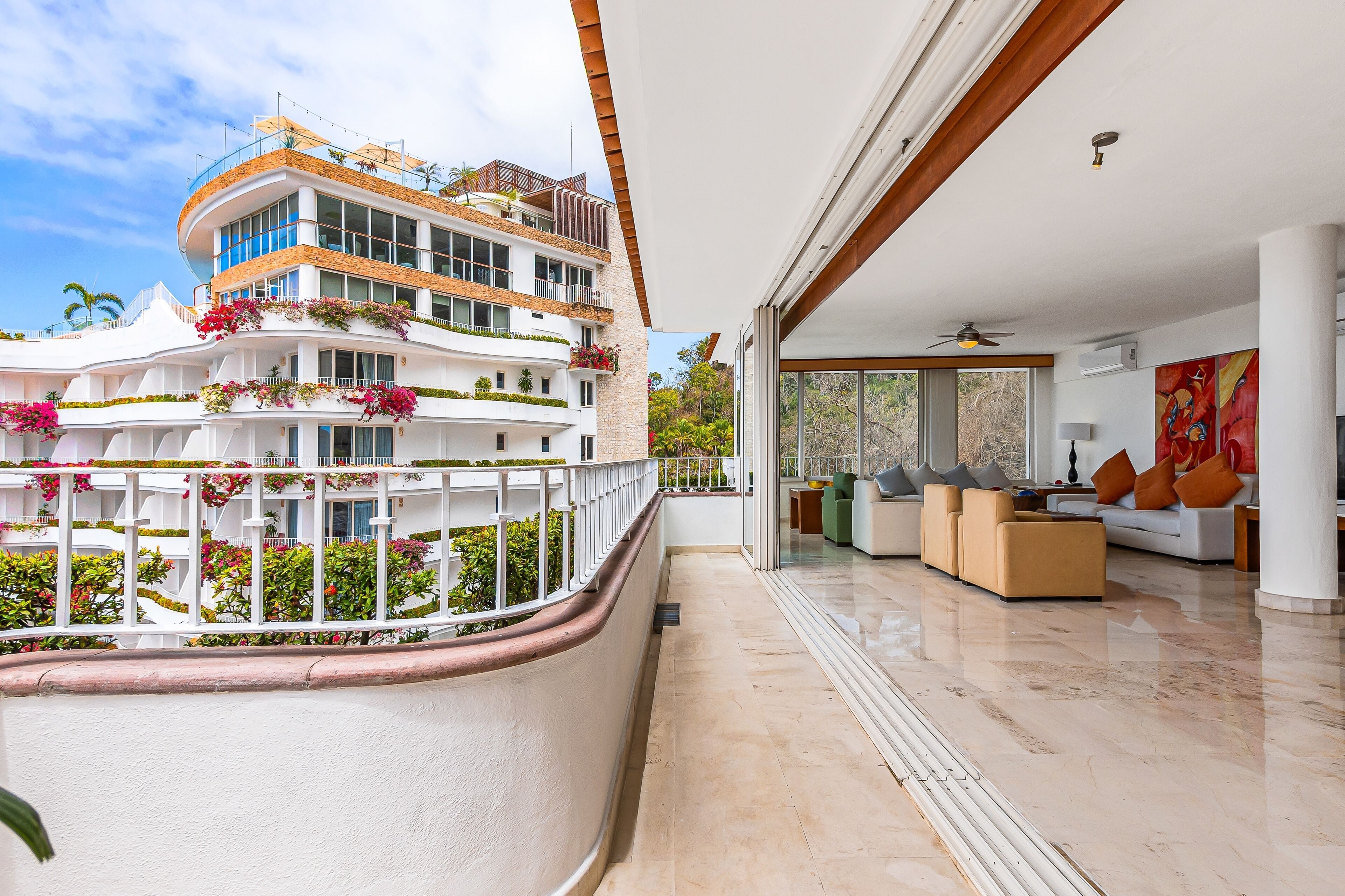 Un hotel moderno con un balcón curvo, un amplio salón con sofás y una mesa de centro, y vistas de un edificio de varios pisos con balcones y plantas.