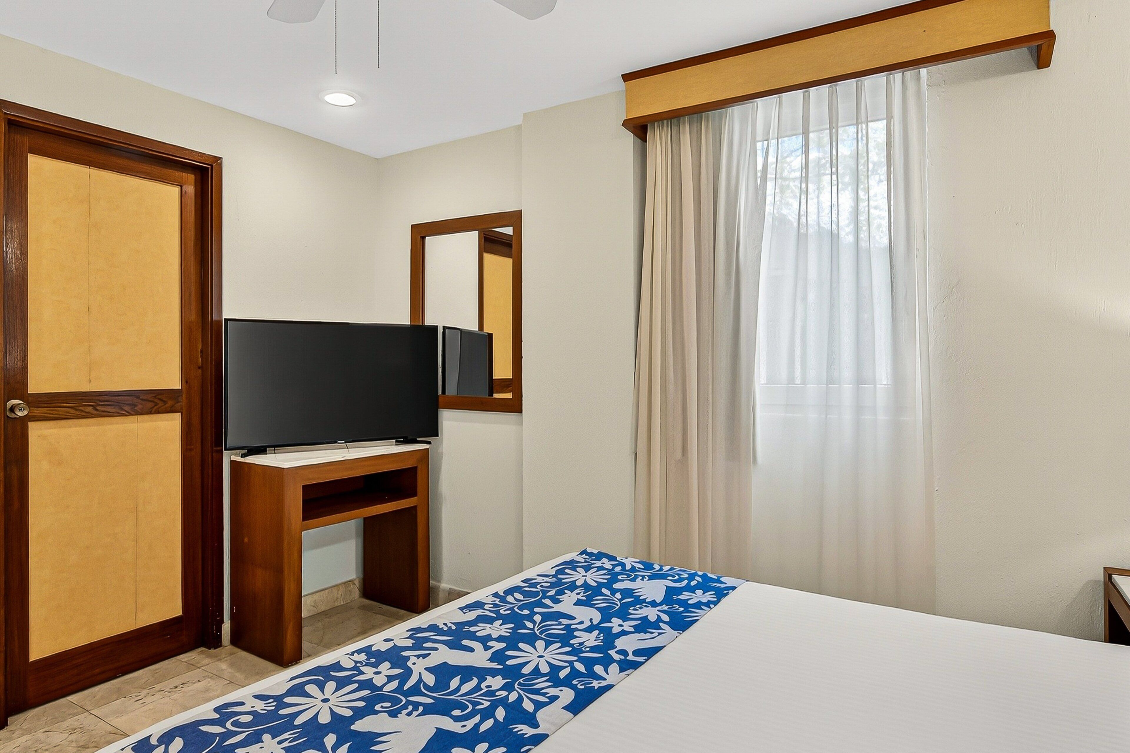 A hotel room with a bed, a TV on a wooden stand, a mirror, and a window with curtains.