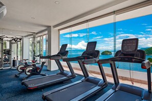 A gym with treadmills, stationary bikes, and weight machines, overlooking a scenic view of the sea. - Stunning Oceanfront Apartment With Breathtaking Views and Luxurious Amenities (Puerto Vallarta)