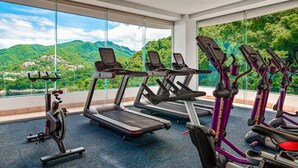 A gym with treadmills, elliptical machines, and a stationary bike, with a view of green hills and a small village through large windows. - Exceptional Waterfront Condo With Pool, Balcony, Fitness Area, & W/D (Puerto Vallarta)