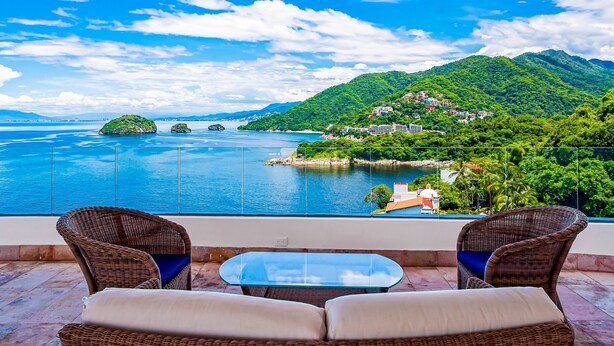 A balcony with wicker furniture, a glass table, and a view of a lake and mountains. - Exceptional Waterfront Condo With Pool, Balcony, Fitness Area, & W/D (Puerto Vallarta)