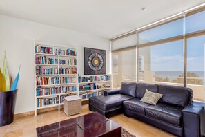 A modern living room with a leather sofa, a bookshelf, and a large window offering a view. - Ocean-view Condo With Pools, Roof Deck, Lounge, Private Balcony, & AC (Nuevo Nayarit)