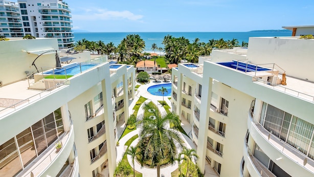 Un hotel de varios pisos con piscina en la azotea, palmeras y vistas al océano.