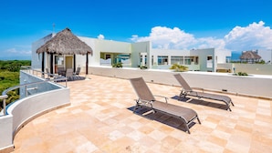 Una terraza en la azotea con sillones de descanso, una estructura con techo de paja y edificios modernos al fondo.