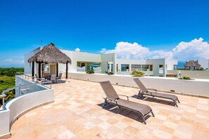 Eine Dachterrasse mit Liegestühlen, einem strohgedeckten Bauwerk und modernen Gebäuden im Hintergrund.