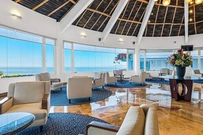 Eine großzügige Lobby mit hoher Decke, großen Fenstern mit Meerblick und einem zentralen Bereich mit Sitzgelegenheiten und einer dekorativen Vase.