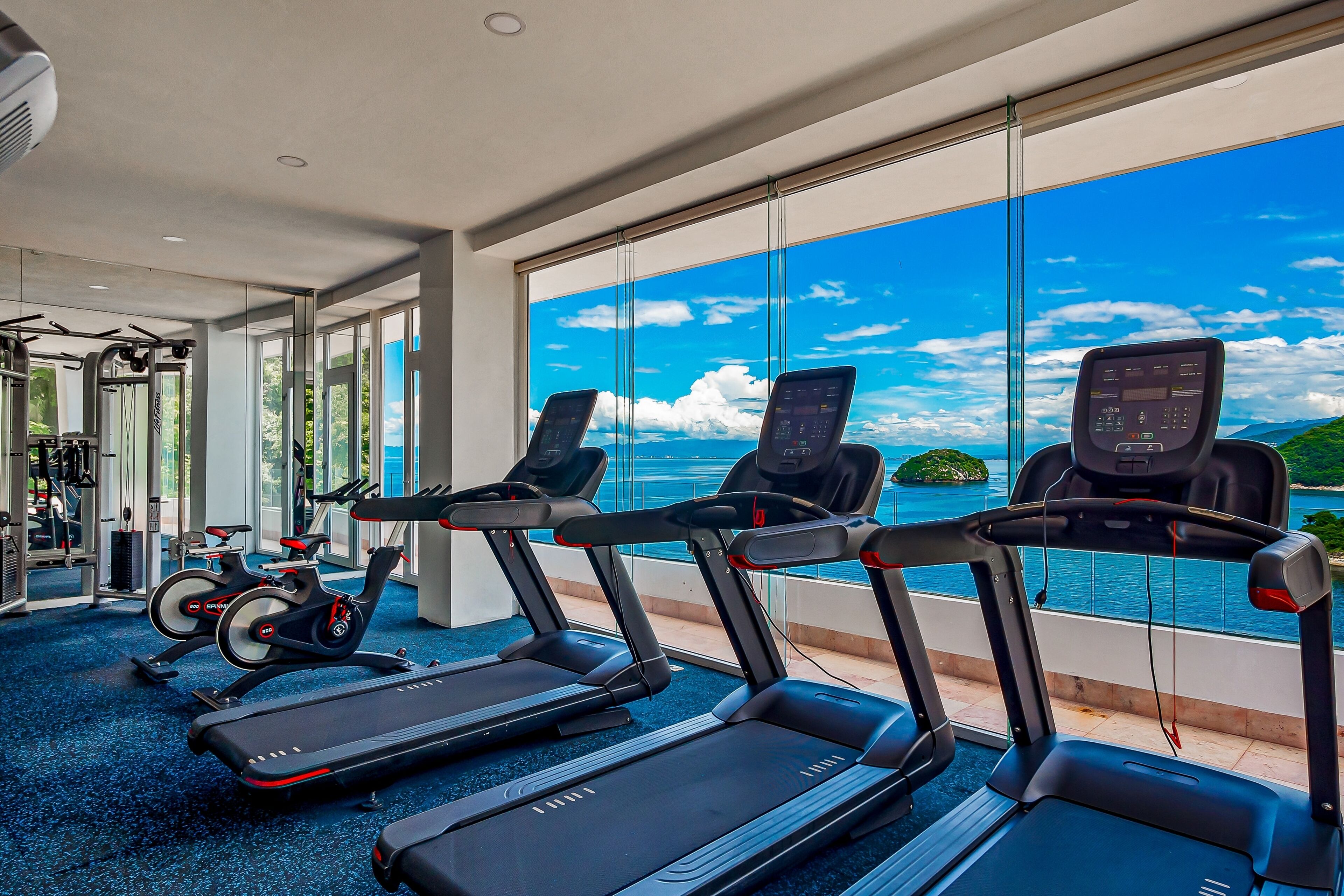 Un gimnasio con cintas de correr, bicicletas estáticas y máquinas de pesas, con vistas panorámicas al mar.