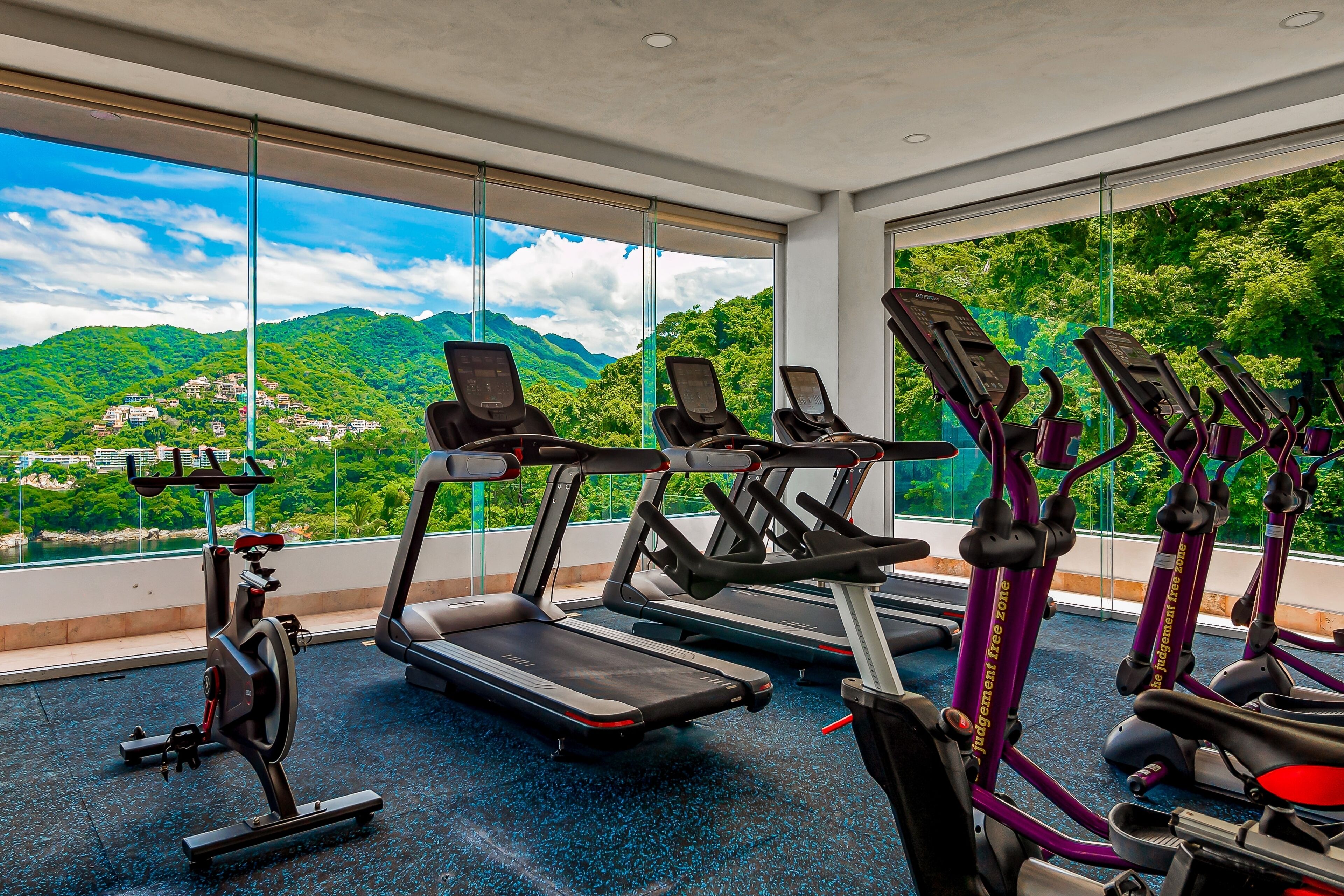 A gym with treadmills, elliptical machines, and a stationary bike, featuring large windows with a mountain view.