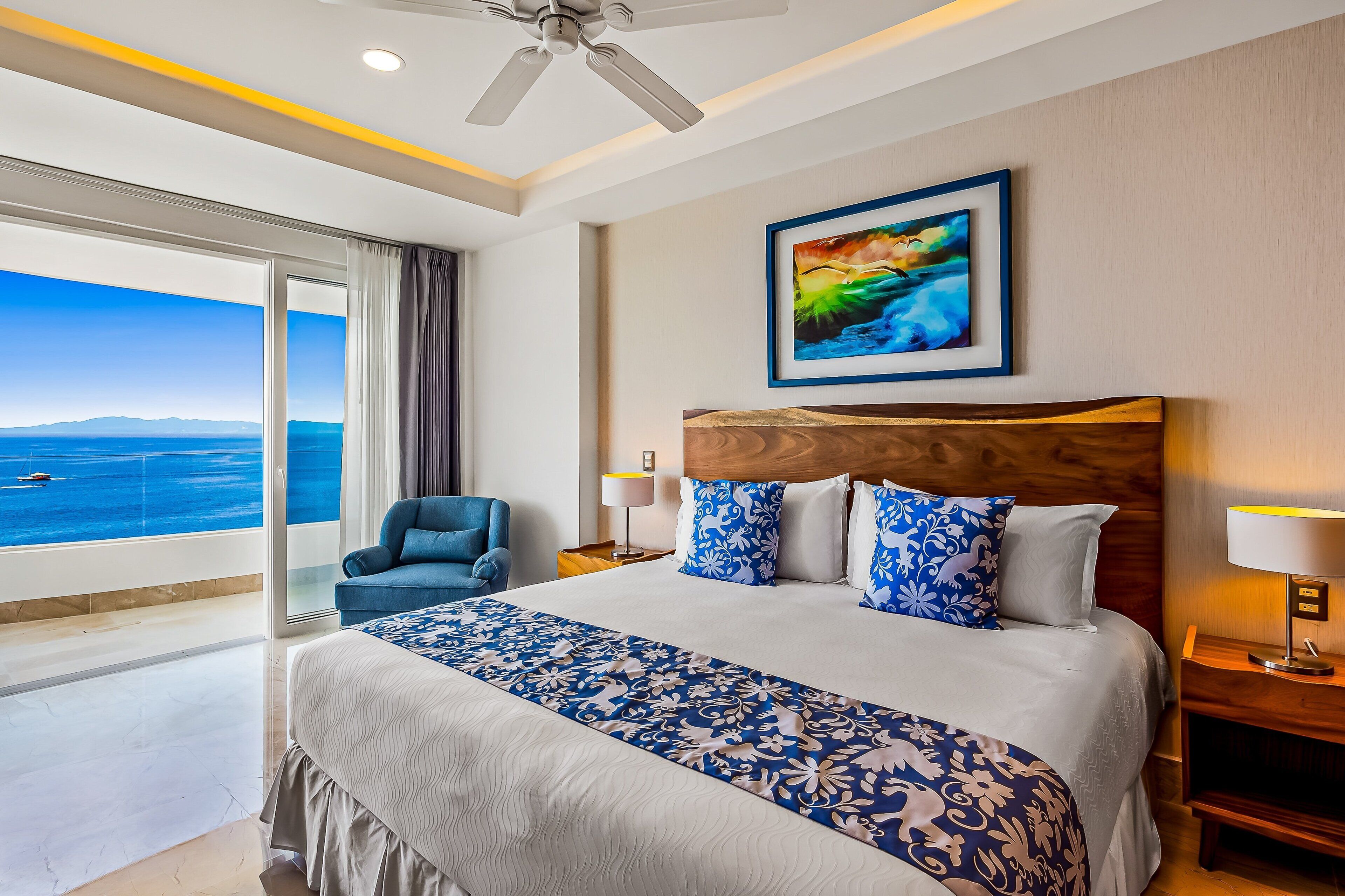A hotel room with a large bed, a view of the sea, a ceiling fan, and a framed painting on the wall.