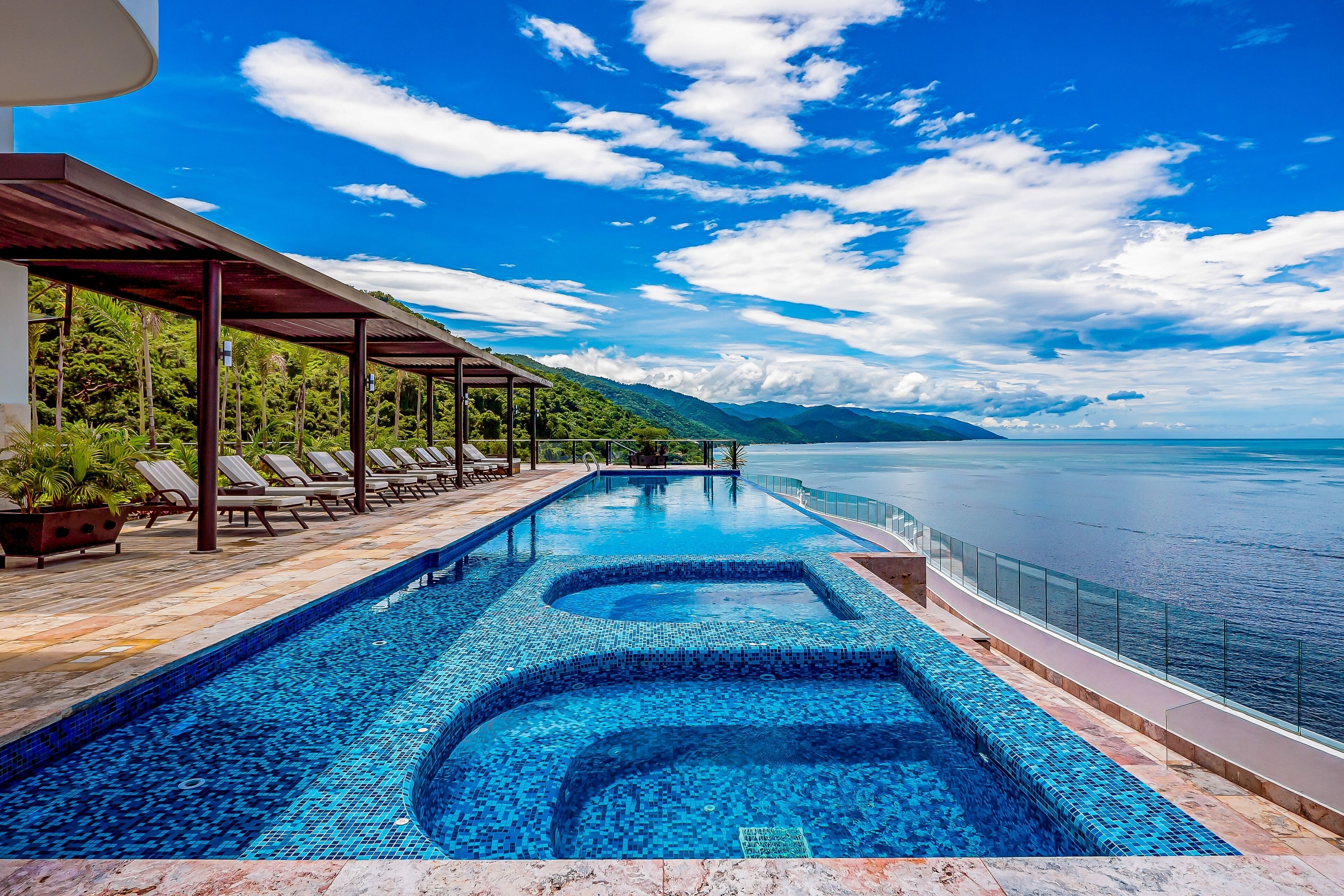 A swimming pool with a tiled edge, a covered seating area with lounge chairs, and a view of the sea and mountains.