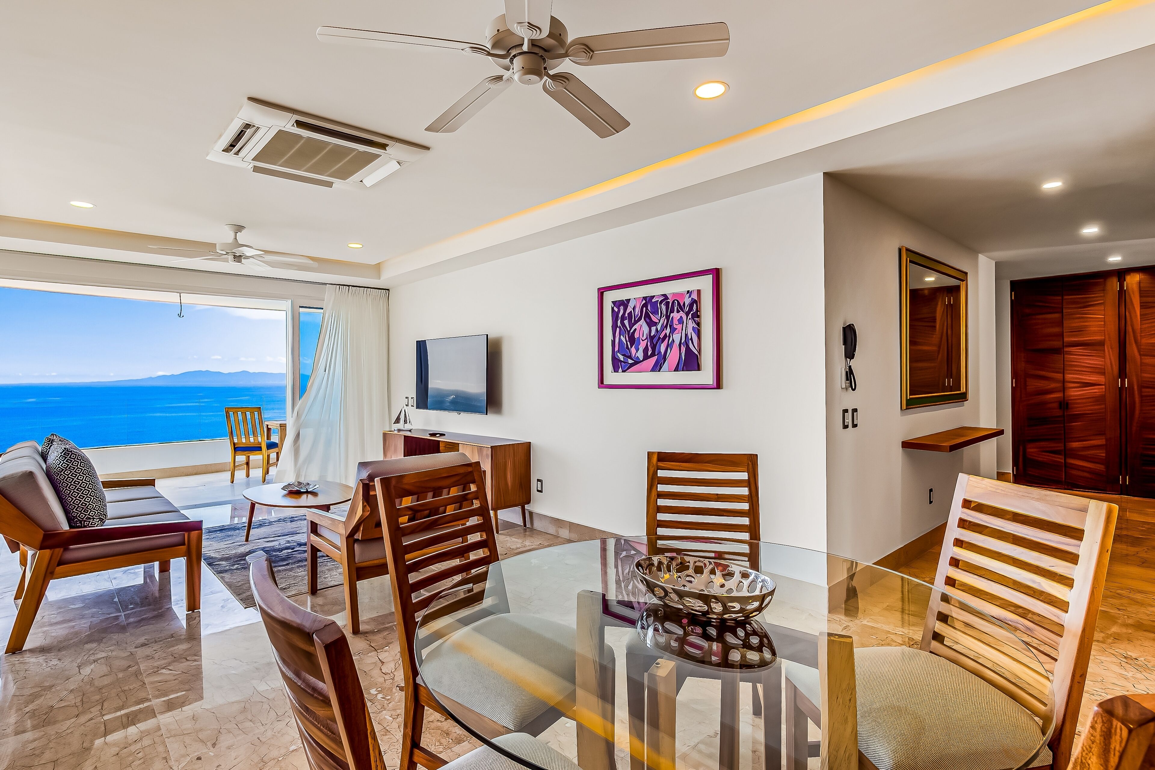 A modern living room with a glass table, wooden chairs, a ceiling fan, and a view of the ocean.