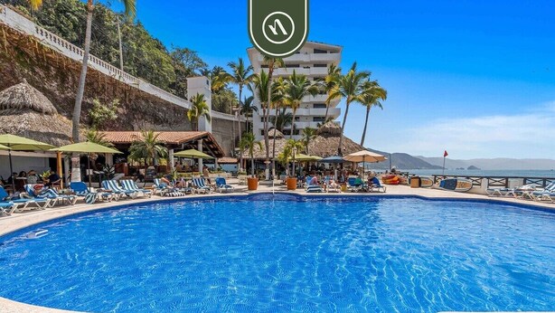 Ein Poolbereich mit Liegestühlen und Sonnenschirmen, umgeben von Palmen und einem Gebäude mit mehreren Balkonen.