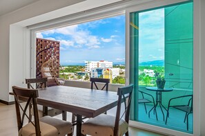 Un comedor con una mesa y sillas, vistas a edificios y vegetación a través de un ventanal grande, y un balcón con mesa y sillas.