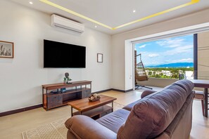 Ein Wohnzimmer mit Flachbildfernseher, Couch, Couchtisch aus Holz und Blick auf eine Stadtkulisse durch ein großes Fenster.