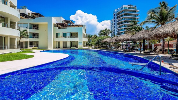 A swimming pool with a curved edge, a thatched-roof structure, and a multi-story building in the background. - Ocean-view Penthouse With Private Plunge Pool & Terrace, Resort Pool (Nuevo Nayarit)