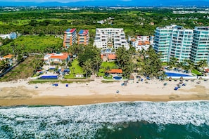 Vista aérea de un hotel frente al mar con varias edificaciones, una playa y palmeras.