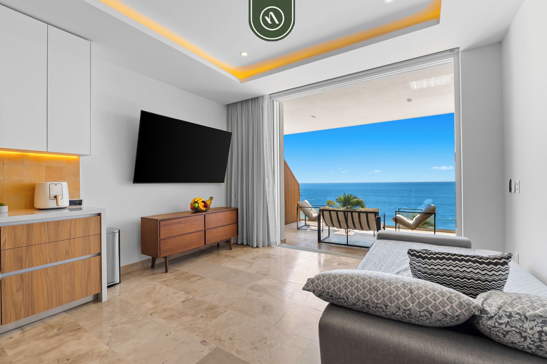 A modern living room with a flat-screen TV, a wooden cabinet, a dining area with a view of the ocean, and a balcony with outdoor seating.