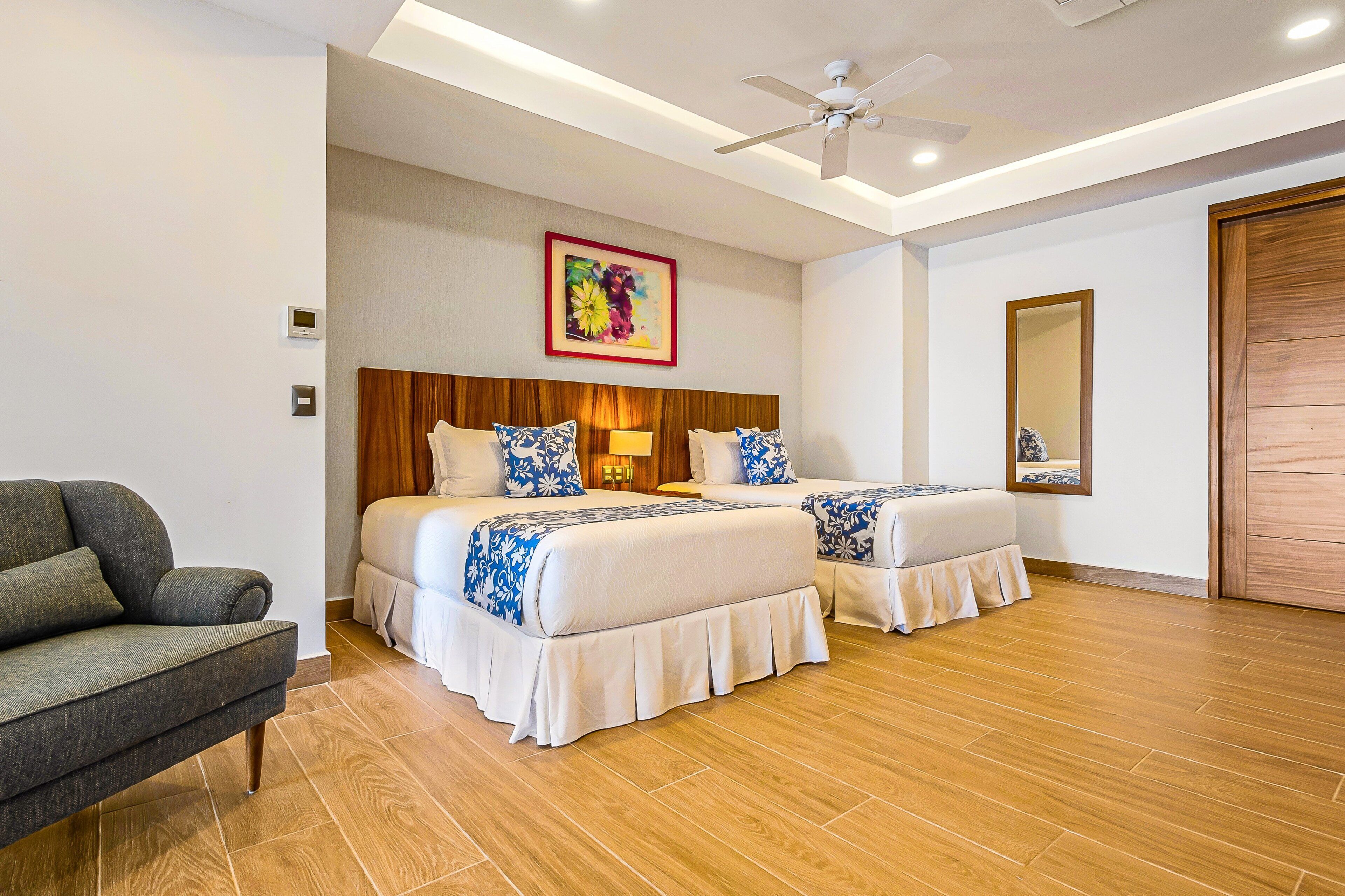 Habitación de hotel con dos camas, un sofá, un ventilador de techo, piso de madera, un cuadro y un espejo.