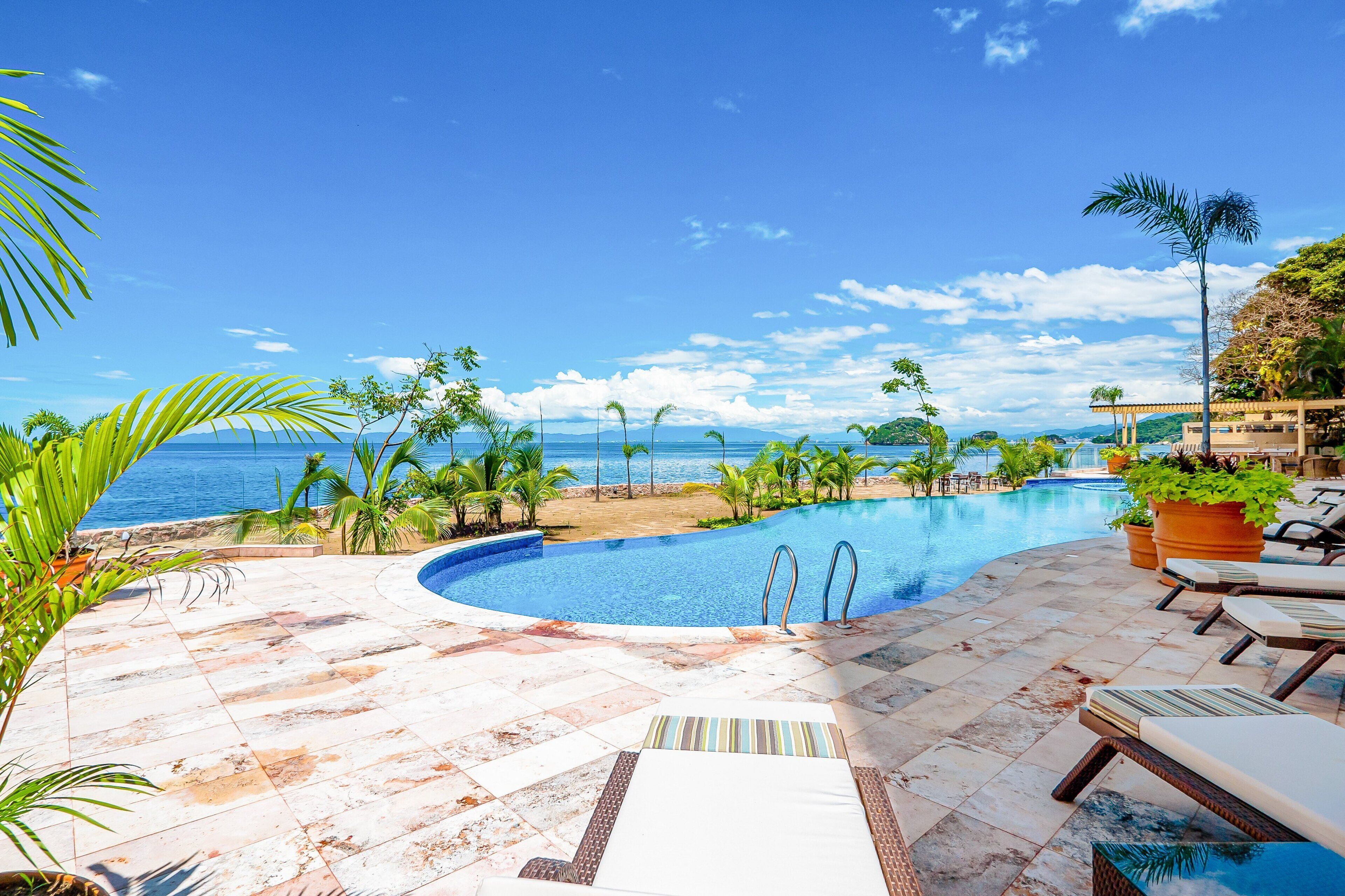 Una piscina con vistas al océano, rodeada de palmeras y sillones de descanso.