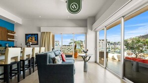 A modern living room with a sofa, dining area, and large windows offering a city view. - Amazing 2 BR Condo in Romantic Zone - Rooftop Pool (Puerto Vallarta)