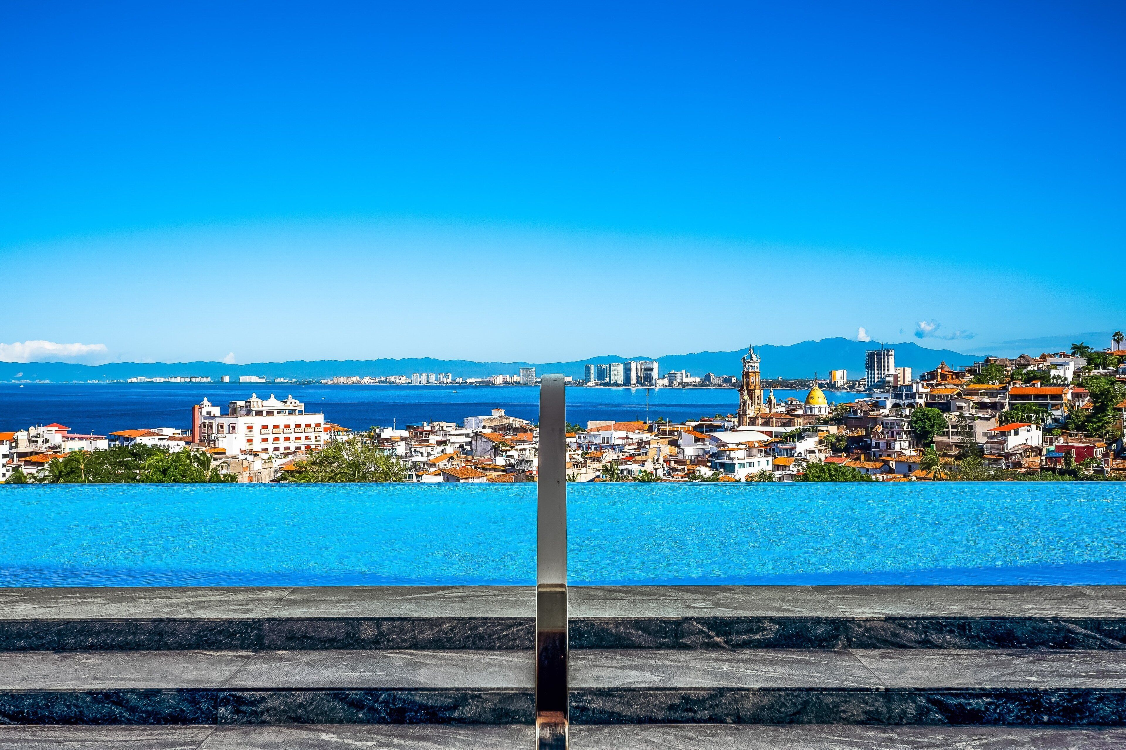 A cityscape with a prominent building in the foreground, a clear blue sky, and a body of water in the background.