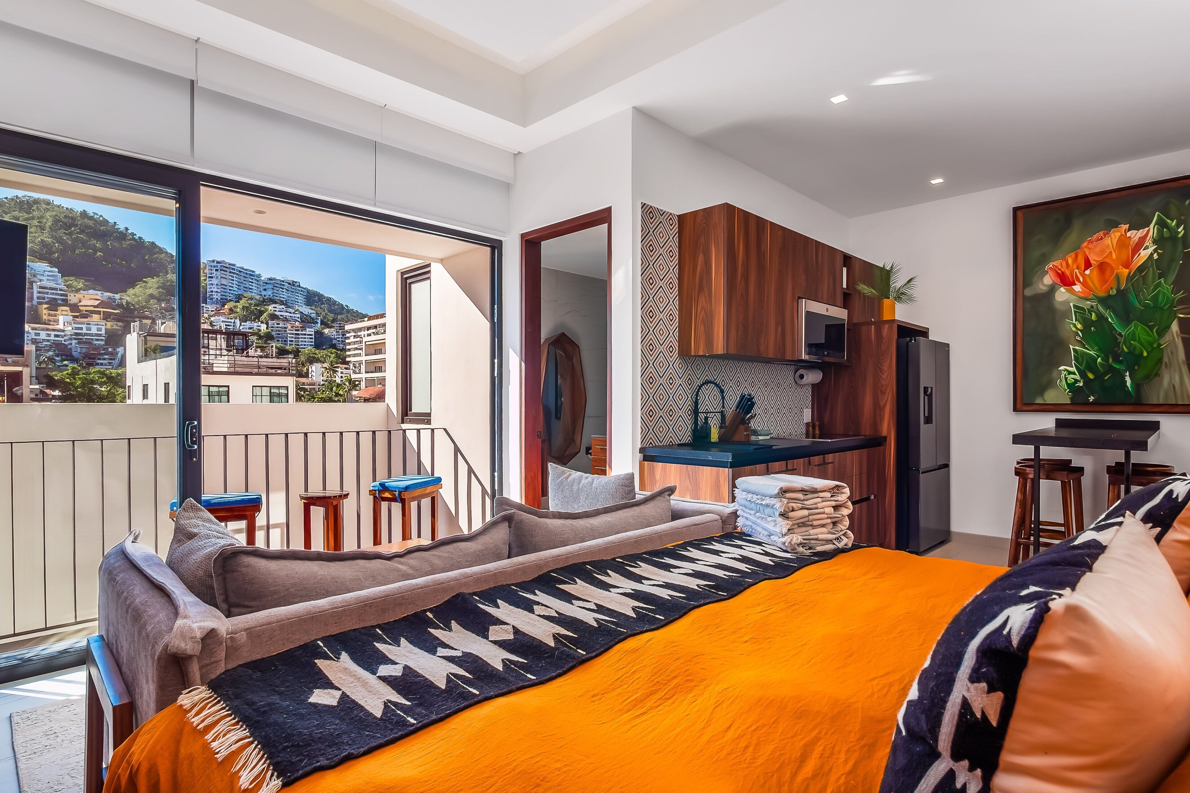 Une chambre d’hôtel moderne avec un balcon, un lit recouvert d’une courtepointe orange, un coin cuisine et une grande fenêtre donnant sur la ville.