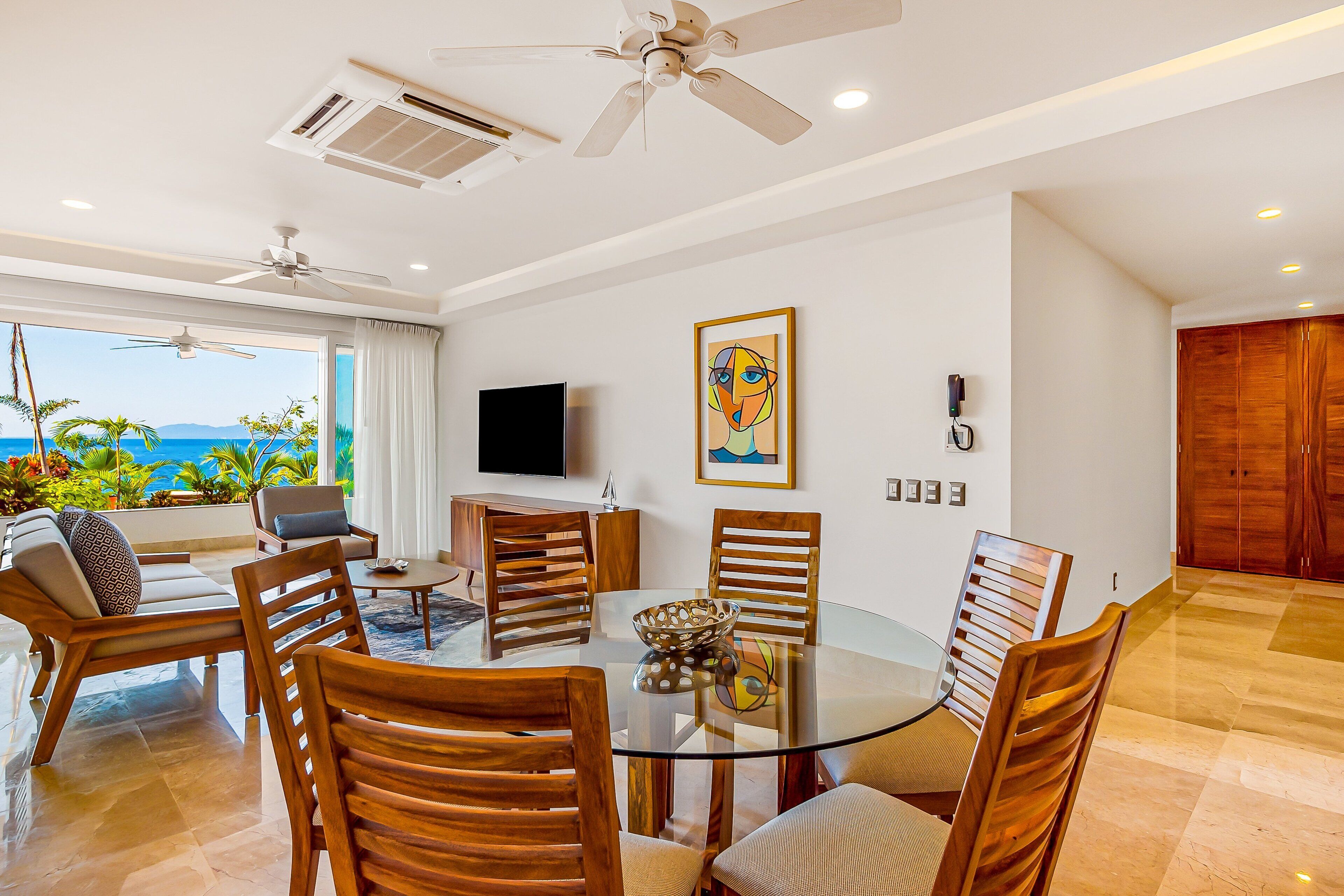 A modern dining area with a glass table, wooden chairs, a TV, and a painting on the wall.