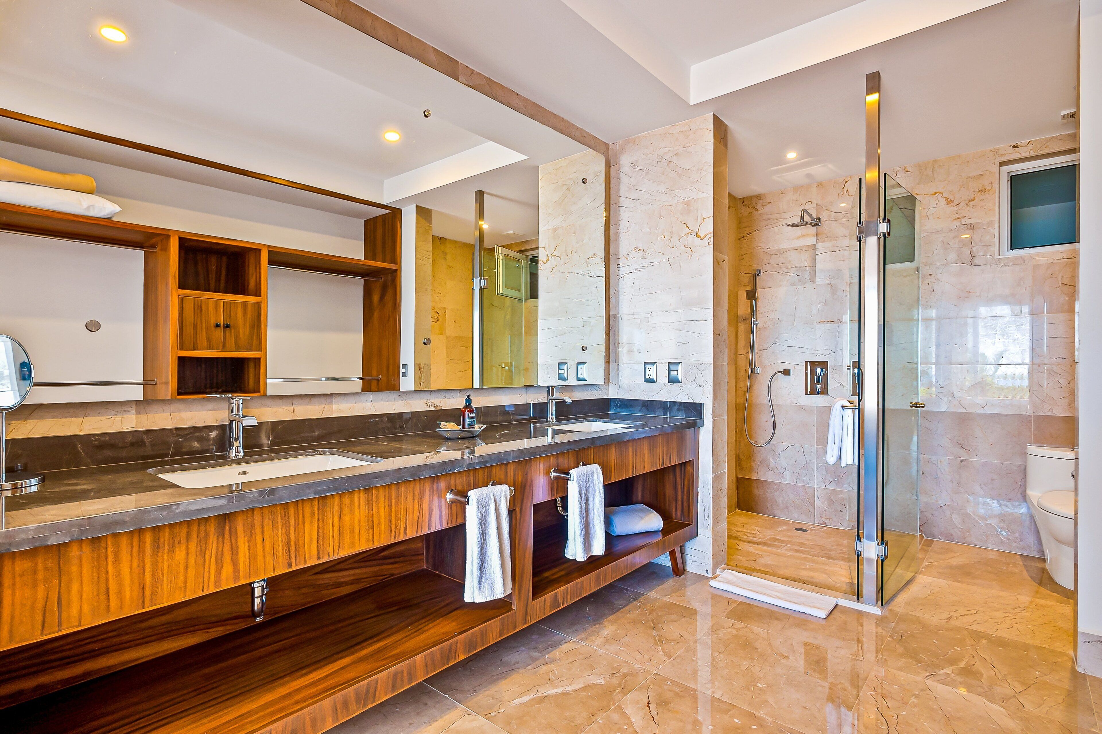Un baño moderno con tocador de madera, lavamanos dobles, un espejo grande y ducha con cerramiento de vidrio.