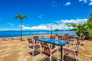 Ett matområde vid stranden med ett glasbord och stolar, palmer och utsikt över havet.