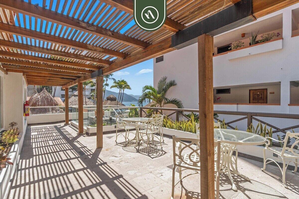 Ein überdachter Außenbereich mit einer Holzpergola, Glastischen und -stühlen sowie Blick aufs Meer.
