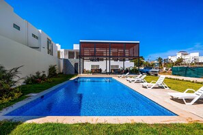 En swimmingpool med liggestole, en moderne bygning med træterrasse, og en klar blå himmel.