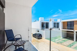 En balkon med gelænder af glas, en blå stol, et rundt bord og en parkeret bil i baggrunden.