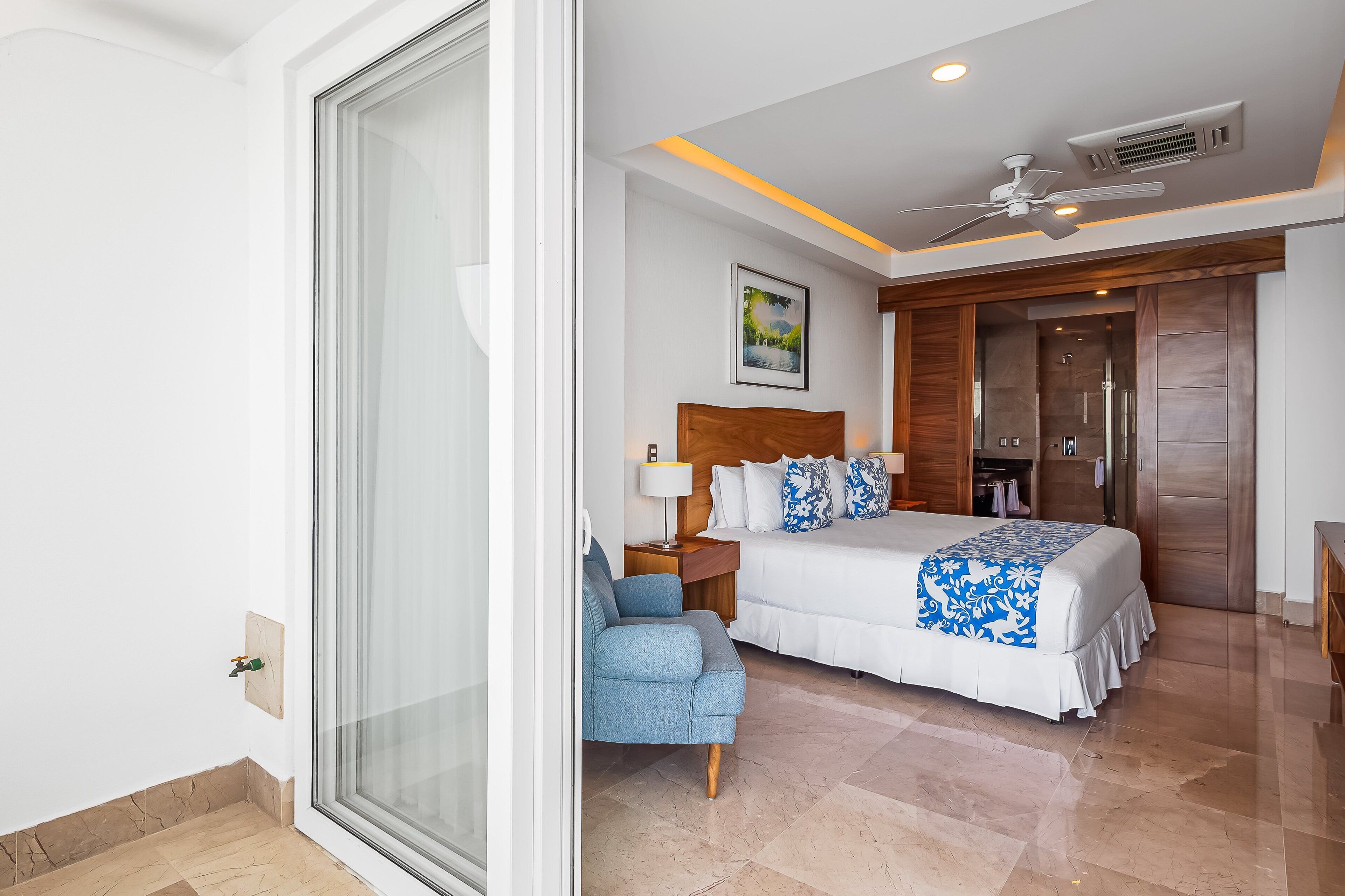 Habitación de hotel con cama, una silla, ventilador de techo y ventana grande.