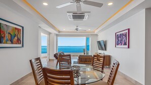 A dining area with a glass table, wooden chairs, ceiling fans, and artwork on the walls. - Luxurious 6th-floor Condo With Pool, Oceanfront Balcony & Water View (Puerto Vallarta)