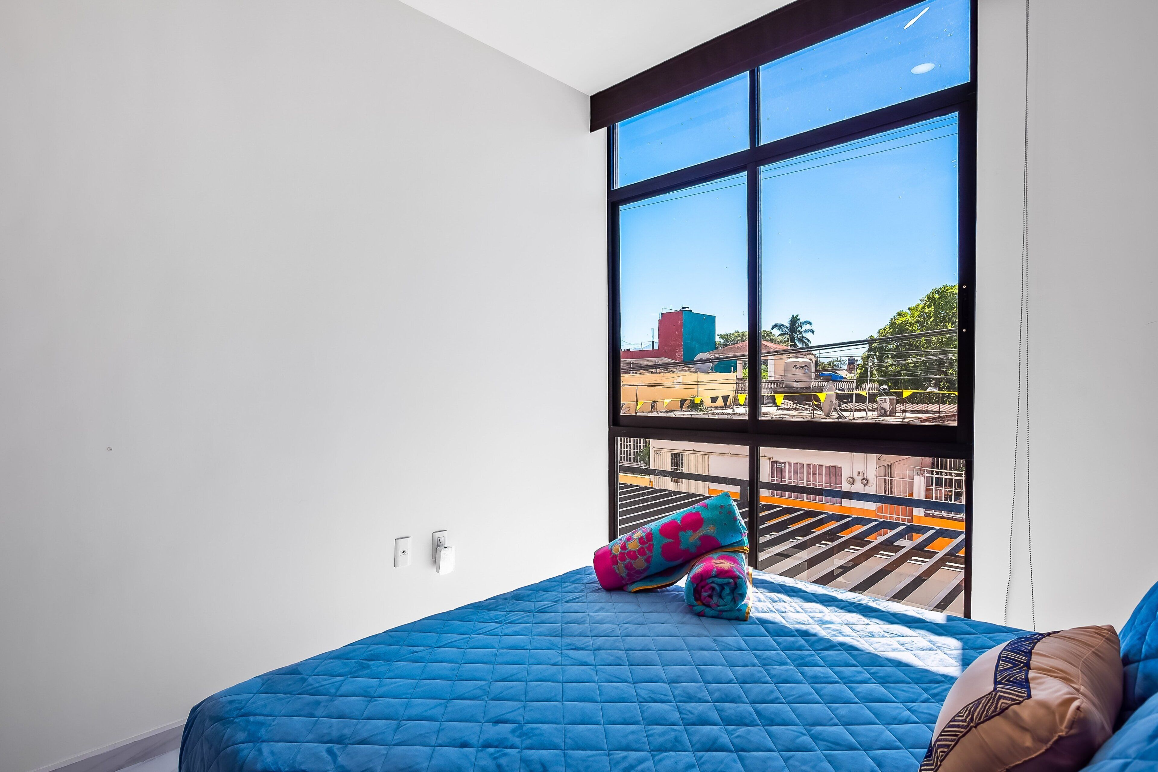 Ein Zimmer mit einem großen Fenster, einem Bett mit blauer Decke und Blick auf eine Stadtlandschaft.