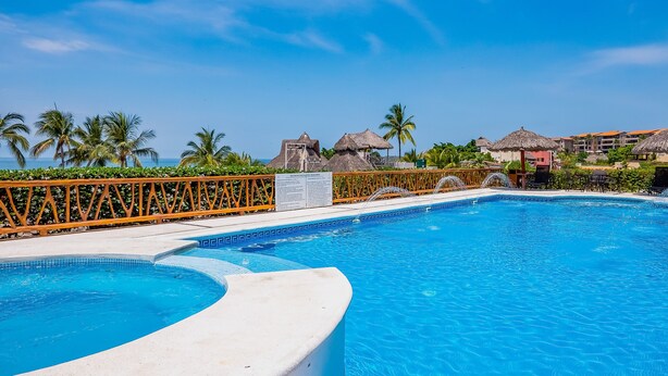 A swimming pool with a curved edge, surrounded by a wooden fence and palm trees. - Stylish Apartment Near Beach & Dining With Pool, hot Tub, Tennis, & Large Patio (Punta de Mita)