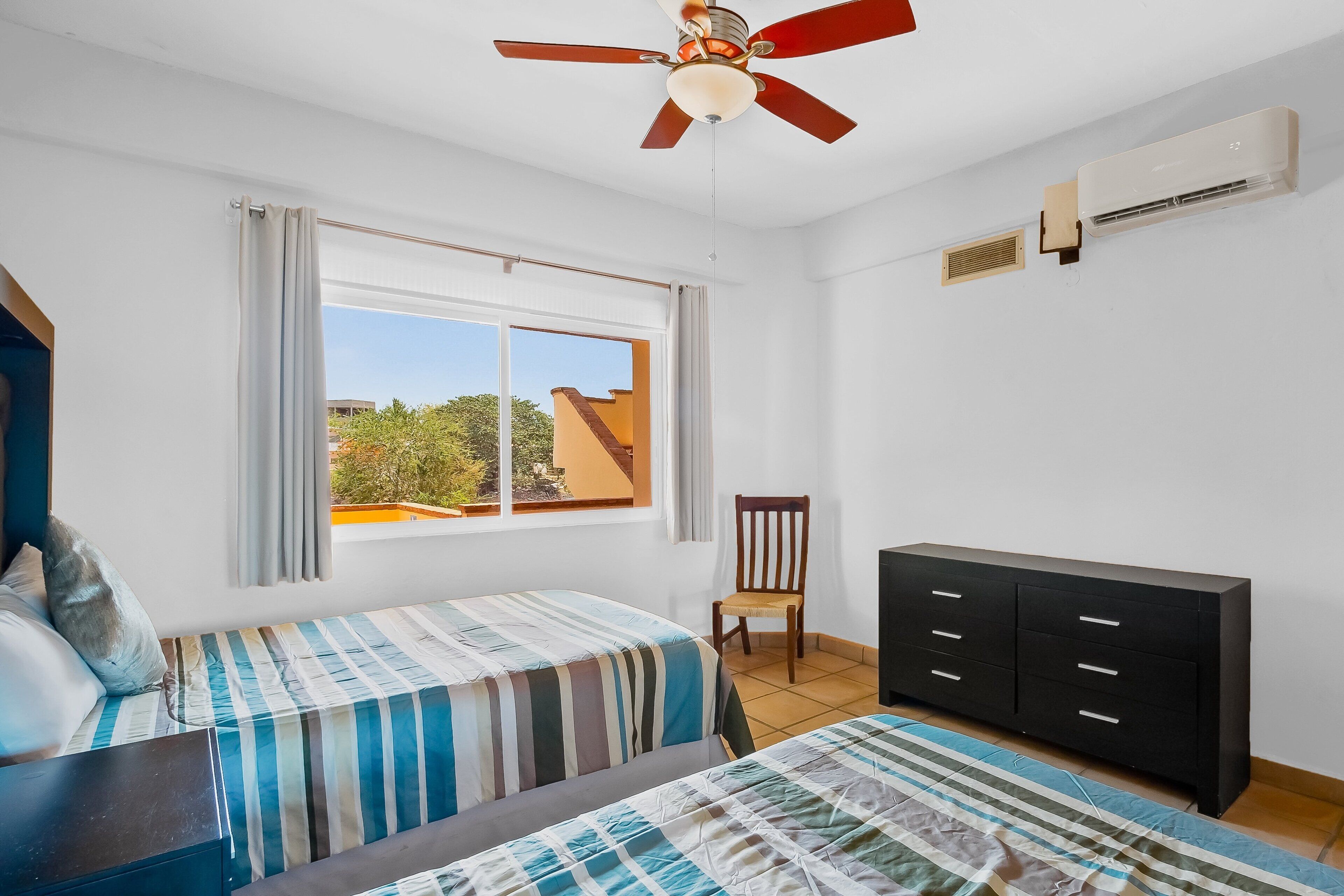 A bedroom with two beds, a ceiling fan, a window with curtains, a chair, a dresser, and an air conditioning unit.