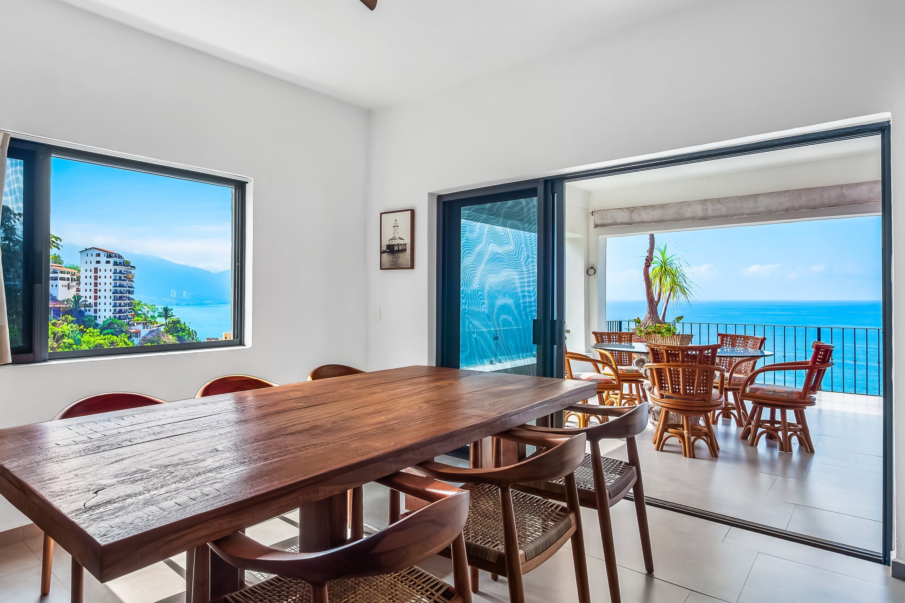 Ein Essbereich mit Holztisch und -stühlen, großen Fenstern mit Blick auf Gebäude und das Meer sowie einem Balkon mit Korbmöbeln.