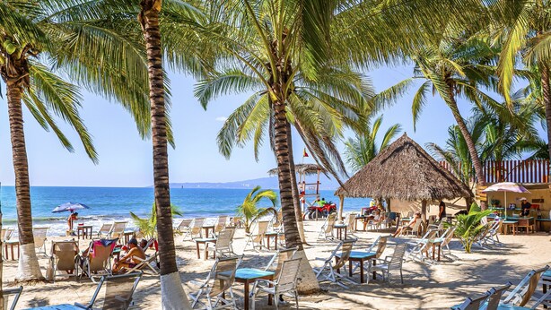 Una playa con palmeras, sillas de descanso y una estructura de techo de paja.