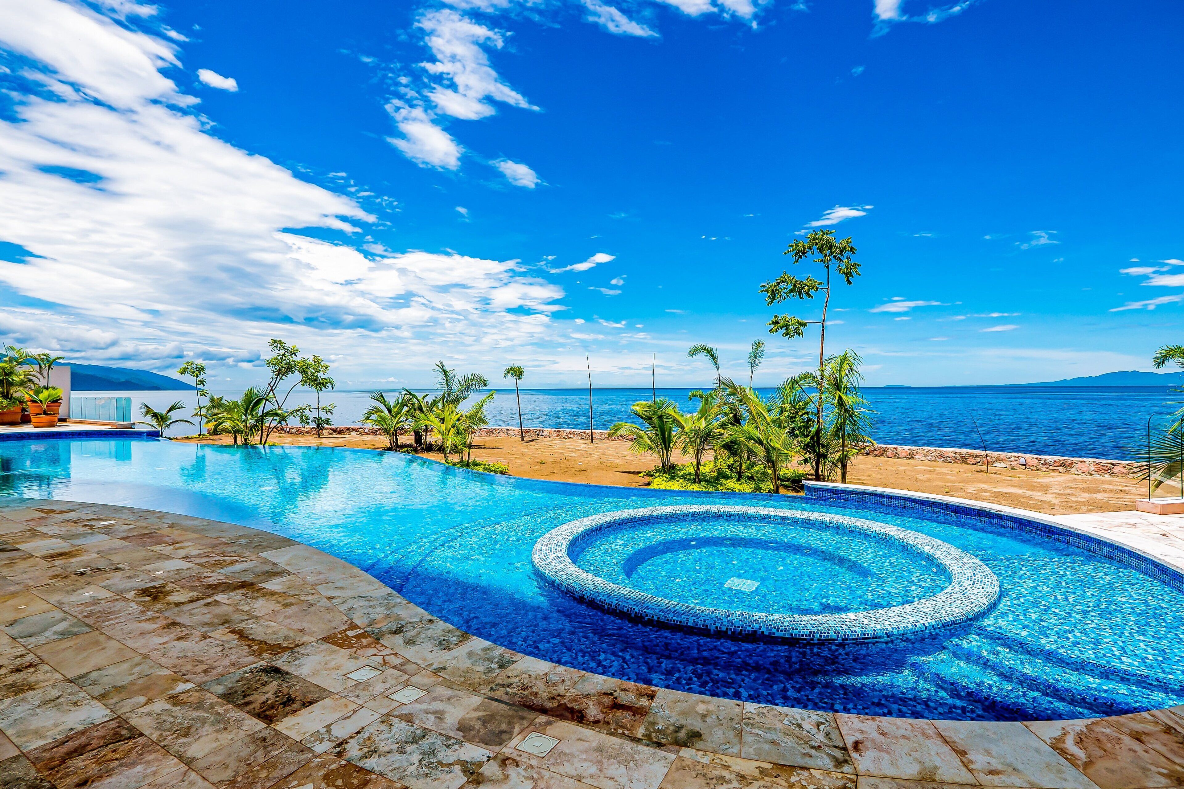 Una piscina con una vasca idromassaggio circolare, circondata da palme e vista sul mare.