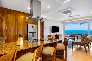 A modern kitchen with a granite countertop, wooden chairs, a built-in stove, and a large window overlooking the ocean. - Fantastic Oceanfront Apartment With Balcony, Pool & Central AC (Puerto Vallarta)