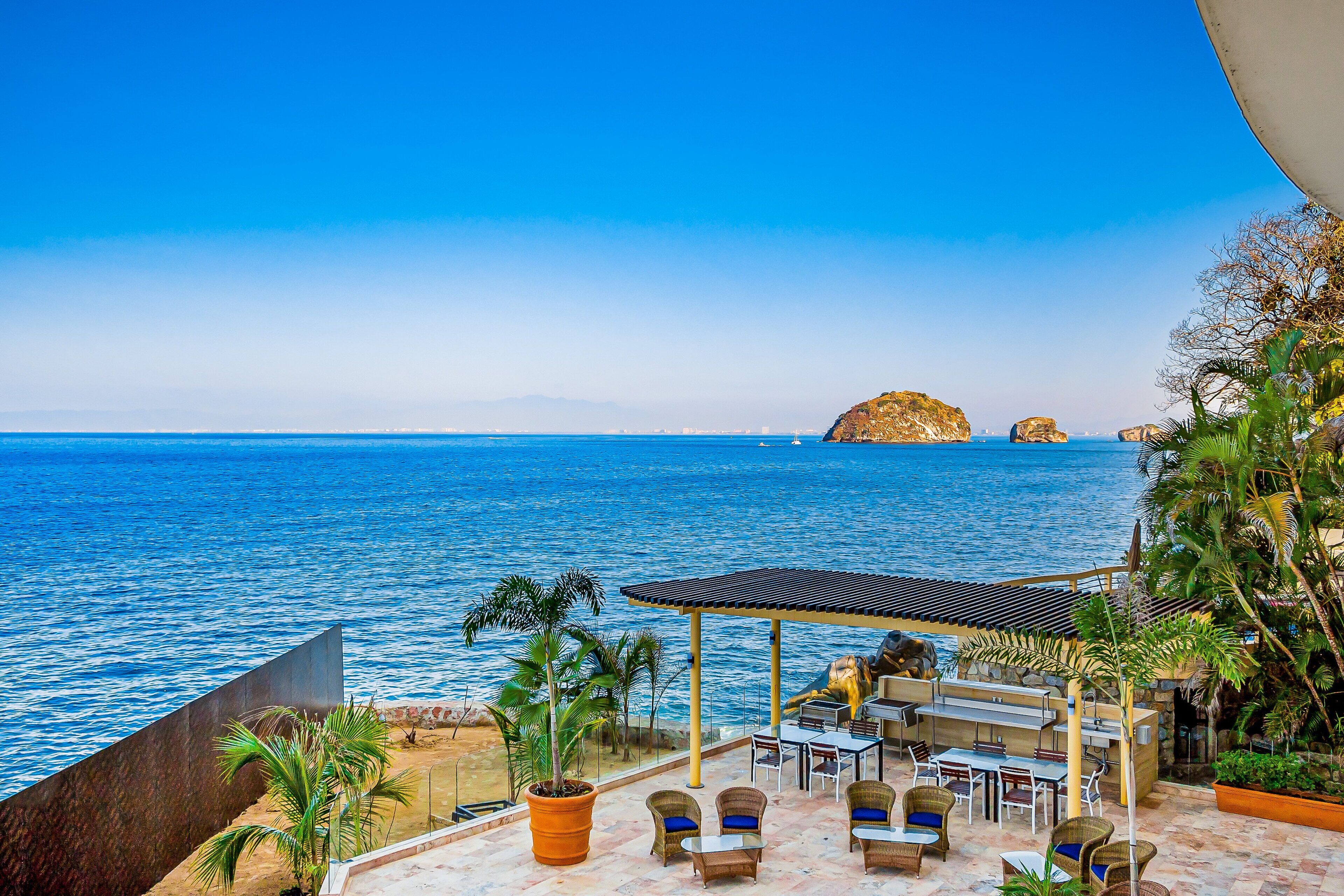 A terrace with outdoor seating overlooking the sea, a palm tree, and a rocky outcrop in the distance.