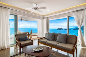 Quarto moderno com sofá, cadeiras, mesa de centro e janelas amplas com vista para o mar.