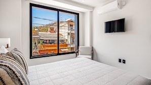 A modern bedroom with a large window, a bed, a chair, a TV, and a wall-mounted air conditioner. - 2nd-floor Condo With Heated Outdoor Pool, Grills, Balcony, W/d, AC (Puerto Vallarta)